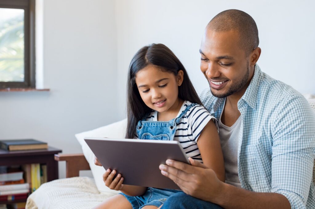 A child and her father smile while watching something on a tablet