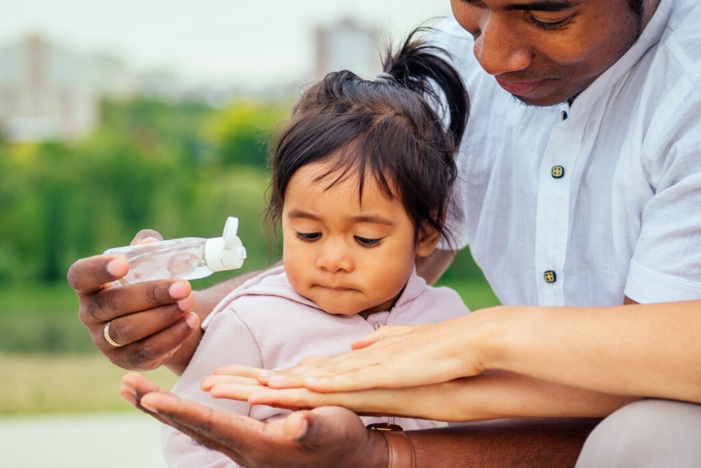 An image of a child with her father helping her put on hand sanitizer outdoors
