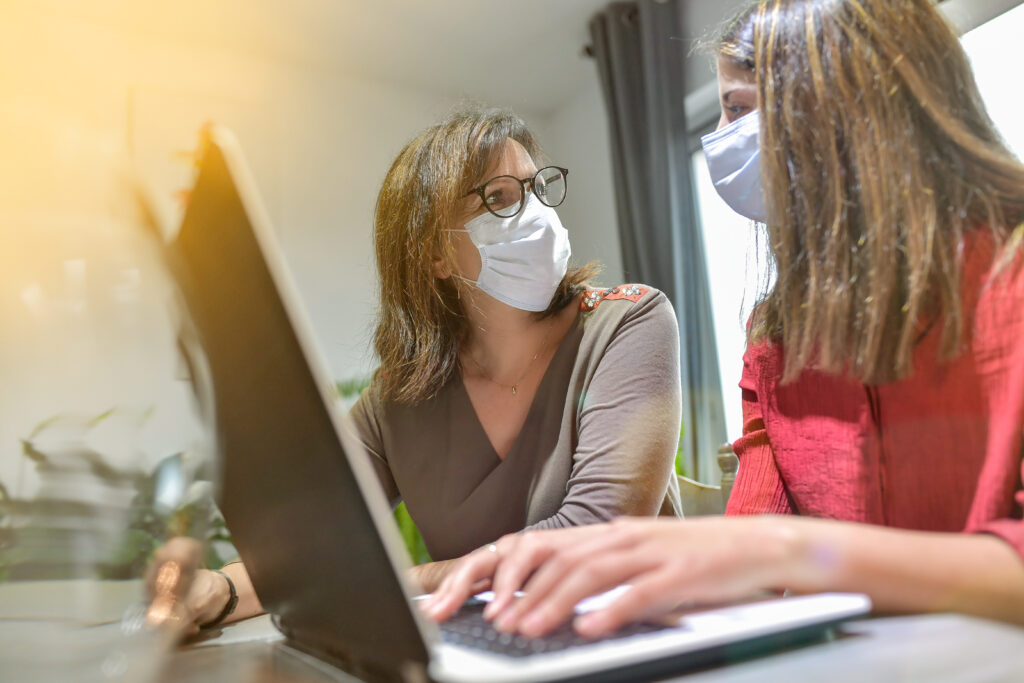 Two individuals are wearing medical masks. They're seated at a table, having a discussion.