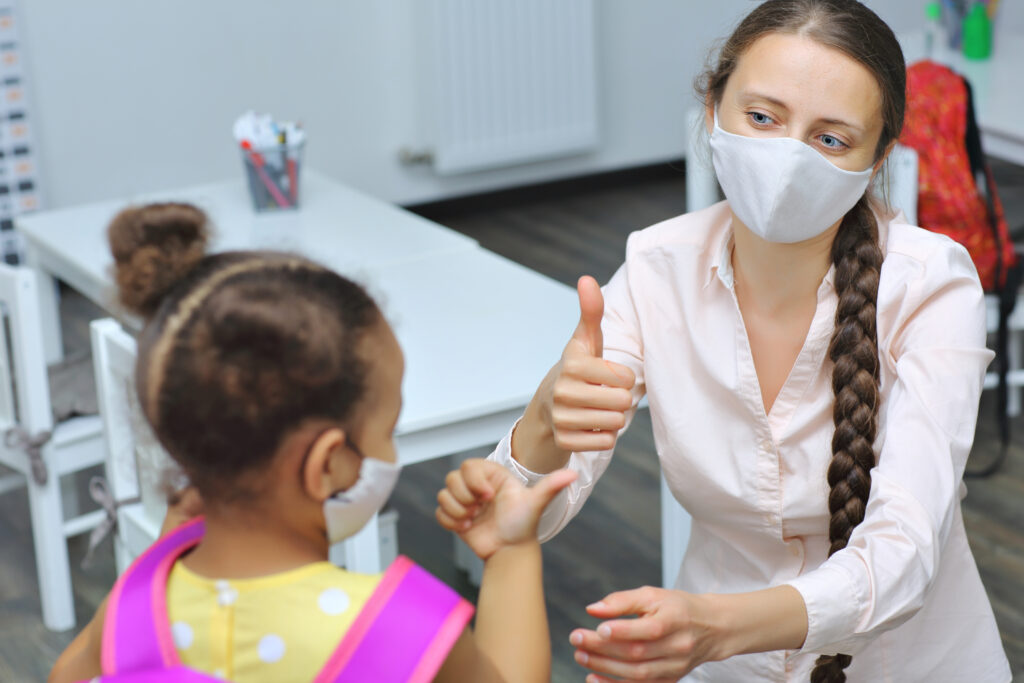 An educator and a child in the learning environment are both wearing masks. They're giving each other the thumbs up.