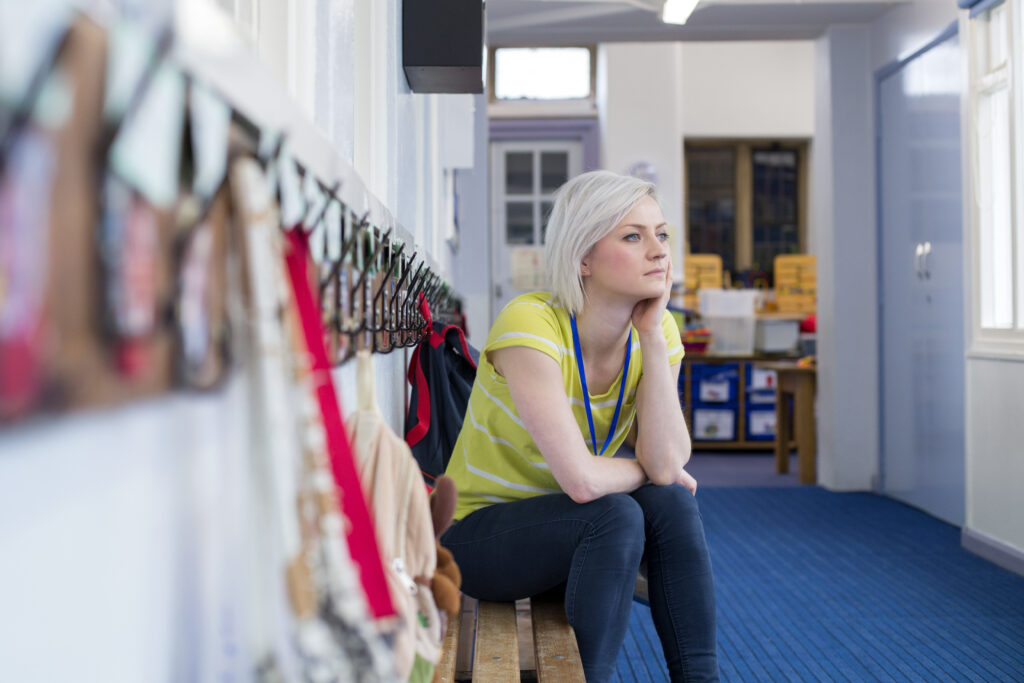 An educator sits in the coat room of a learning environment. She looks visibly pensive.