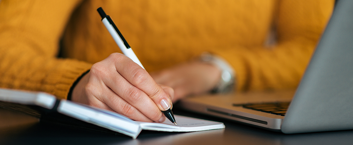 Image d’une personne prenant des notes sur un cahier avec son ordinateur devant elle.